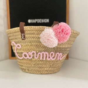 May include: A natural woven straw basket with brown leather handles and pink pom poms. The name "Carmen" is embroidered on the front of the basket in pink thread.