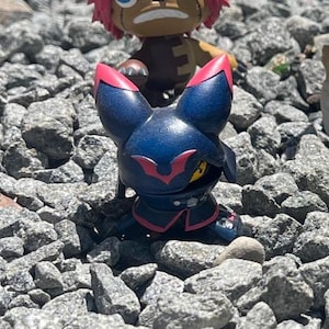 May include: A small, stylised figurine featuring a dark blue, cat-like character with pointed ears, red accents, and a visible yellow eye. The figure has a rounded head and a compact body, positioned in front of a partially visible brown character.