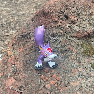 Peut inclure: Figurine violette et blanche représentant une créature avec un œil rouge et des griffes noires. La figurine est posée sur une roche brun-rouge, avec un arrière-plan flou d'éléments naturels.