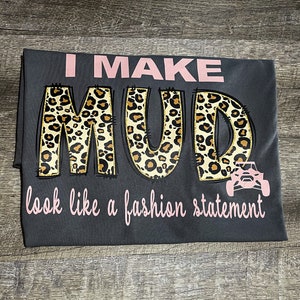 May include: Gray t-shirt with the text "I MAKE MUD look like a fashion statement" in pink lettering. The word "MUD" is in leopard print with a small pink ATV graphic.