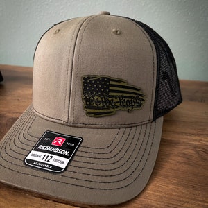 May include: A brown and black trucker hat with a distressed American flag patch that says "We The People". The hat has a black mesh back and a Richardson 112 adjustable label.