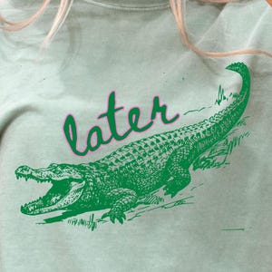 May include: A light green t-shirt with a green alligator graphic and the word "later" in a script font. The alligator is detailed with open jaws. The shirt is a relaxed fit.