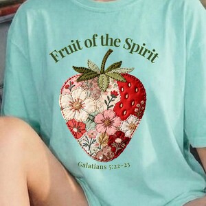 May include: A light blue t-shirt with the text "Fruit of the Spirit" above a strawberry design. The strawberry is red with a floral pattern and green leaves. The text "Galatians 5:22-23" is below the strawberry.