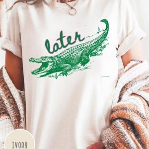 May include: Ivory-colored t-shirt featuring a green alligator graphic with the word "later" above it. The alligator is detailed with scales and open jaws. The shirt has rolled-up sleeves and a casual fit.