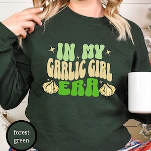 May include: A forest green long-sleeve sweatshirt featuring the text "IN MY GARLIC GIRL ERA" in a retro font. The lettering is in light green and cream, accompanied by two graphic illustrations of garlic bulbs on either side of the word "ERA".