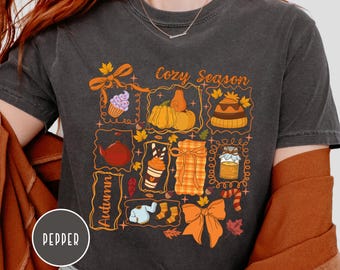May include: Dark gray t-shirt with a fall-themed graphic. The design includes a cupcake, pumpkins, a hat, a teapot, a latte, a wrapped gift, a jar of jam, socks, and a bow. The words "Cozy Season" and "Autumn" are also featured.