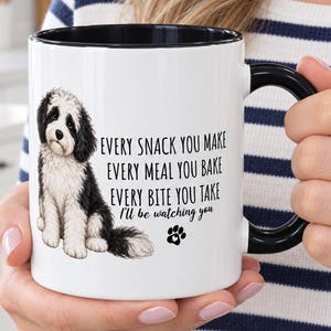 May include: A white ceramic mug with a black handle and interior rim. The mug features a black and white illustration of a fluffy dog sitting down, accompanied by the text: Every snack you make, every meal you bake, every bite you take, I'll be watching you, and a small black paw print icon.