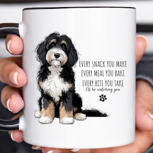 May include: White ceramic mug with a black handle and rim, featuring a black, brown, and white dog illustration. Text reads: "EVERY SNACK YOU MAKE, EVERY MEAL YOU BAKE, EVERY BITE YOU TAKE, I'll be watching you."