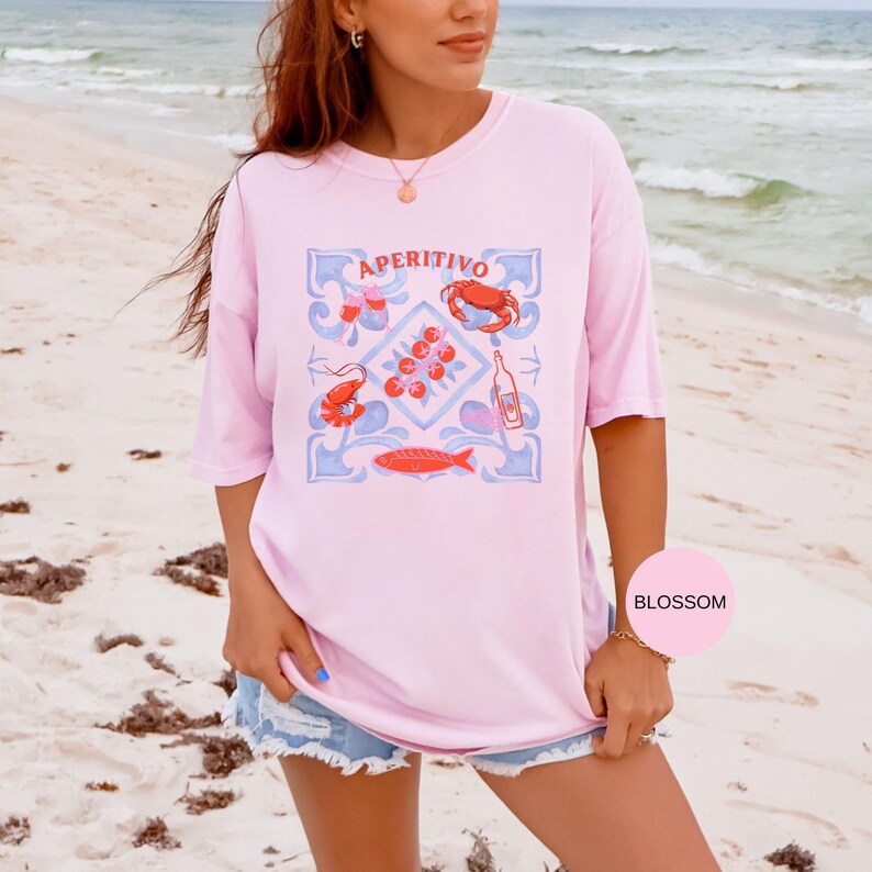 Op de afbeelding: Een lichtroze t-shirt met het woord "APERITIVO" boven een blauw en rood grafisch ontwerp van zeevruchten en een fles. Het shirt wordt gedragen met een spijkerbroekshort op een strand.