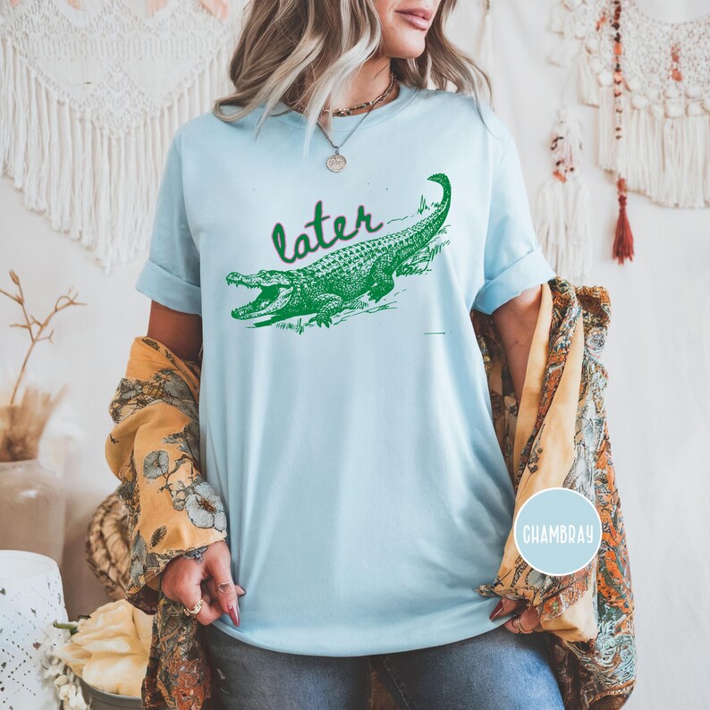 May include: Light blue t-shirt with a green alligator graphic and the word "later" in a cursive font. The alligator is detailed with scales and open jaws. The shirt has rolled-up sleeves.