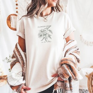 May include: Cream-colored t-shirt featuring a floral illustration with the text "FLOWERS GROW in the Valley" in black. The shirt has rolled-up sleeves. A patterned sweater is draped over the shoulders.