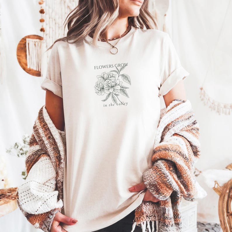 May include: Cream-colored t-shirt featuring a floral illustration with the text "FLOWERS GROW in the Valley" in black. The shirt has rolled-up sleeves. A patterned sweater is draped over the shoulders.