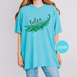 May include: A turquoise t-shirt with a green alligator graphic and the word "later" in cursive. The alligator is detailed with scales and open jaws. The shirt has a relaxed fit and a V-neck.