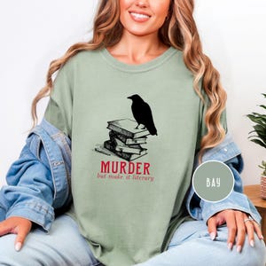 May include: Sage green t-shirt featuring a black raven perched atop a stack of books. The text "MURDER but make it literary" is printed in red below the books. The shirt has a relaxed fit.