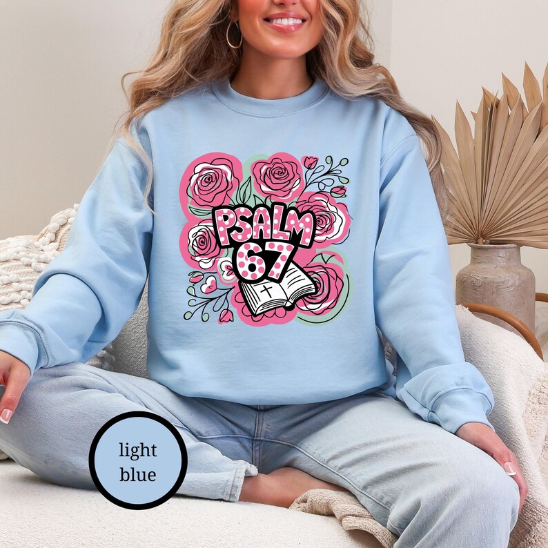 May include: Light blue sweatshirt featuring a floral design with pink roses and the text "PSALM 67". The design includes an open book and is surrounded by green vines. The sweatshirt is a solid light blue color.