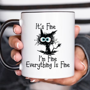 May include: White ceramic mug with a black handle. The mug features a black and white cartoon cat with blue eyes and the text "It's Fine I'm Fine Everything Is Fine."