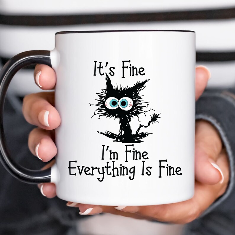 May include: White ceramic mug with a black handle. The mug features a black and white cartoon cat with blue eyes and the text "It's Fine I'm Fine Everything Is Fine."