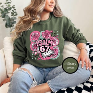 May include: Military green sweatshirt with a floral design featuring pink roses and the text "PSALM 67". The design includes an open book with a cross. The sweatshirt is a crew neck style.