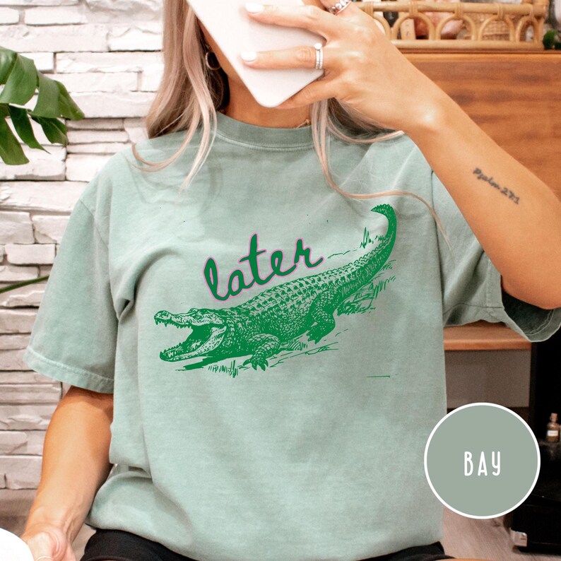 May include: A light green t-shirt with a green alligator graphic and the word "later" in a script font. The alligator is detailed with open jaws. The shirt is a relaxed fit.