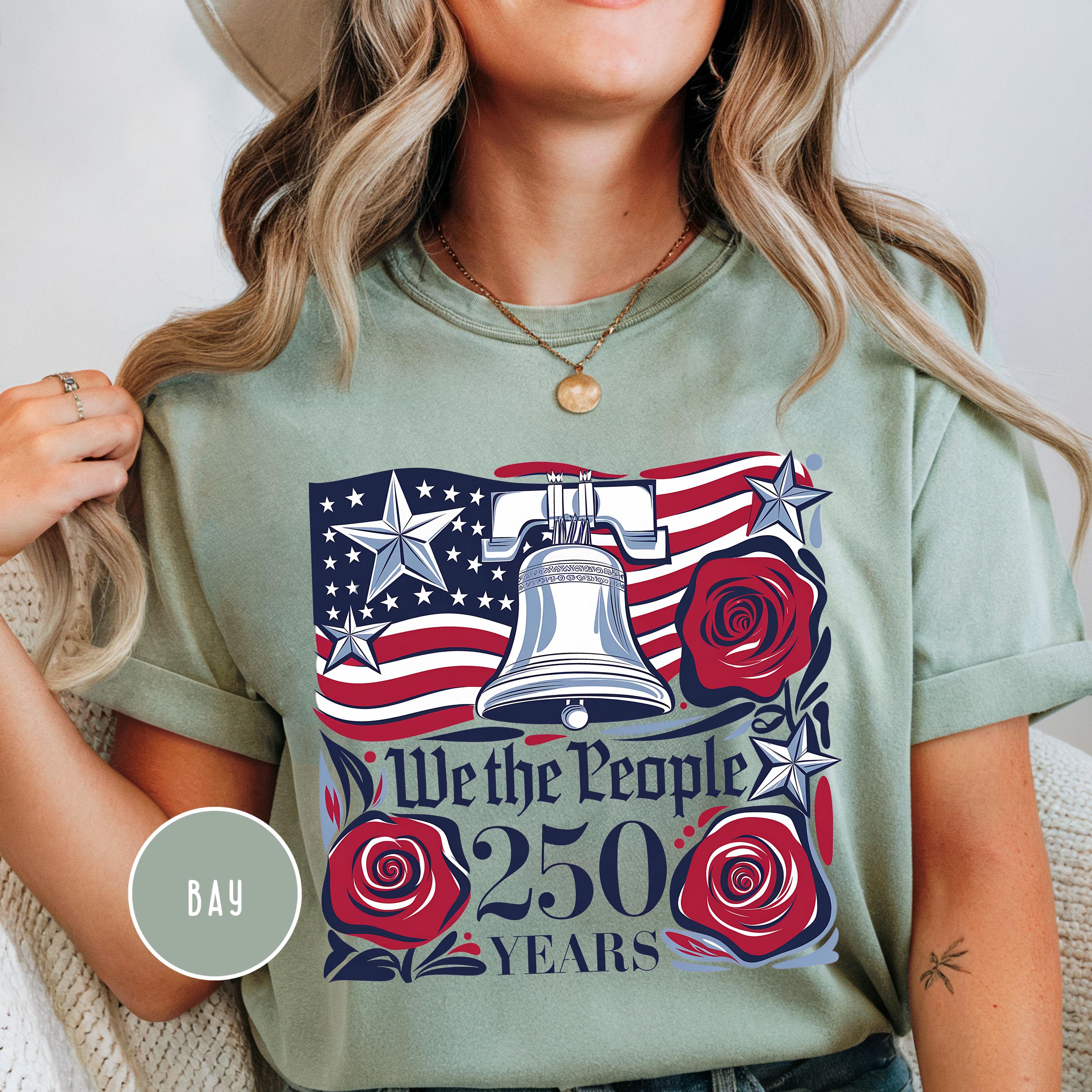 May include: A light green t-shirt with a patriotic design. The design features the American flag, the Liberty Bell, red roses, and the text "We the People 250 Years". The shirt has short sleeves.