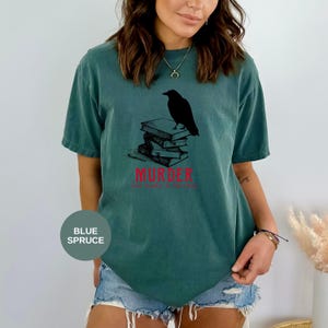 May include: Blue spruce t-shirt featuring a black raven perched atop a stack of books. The word "MURDER" is printed in red, with the phrase "her make a library" below. A casual, comfortable tee for book lovers.