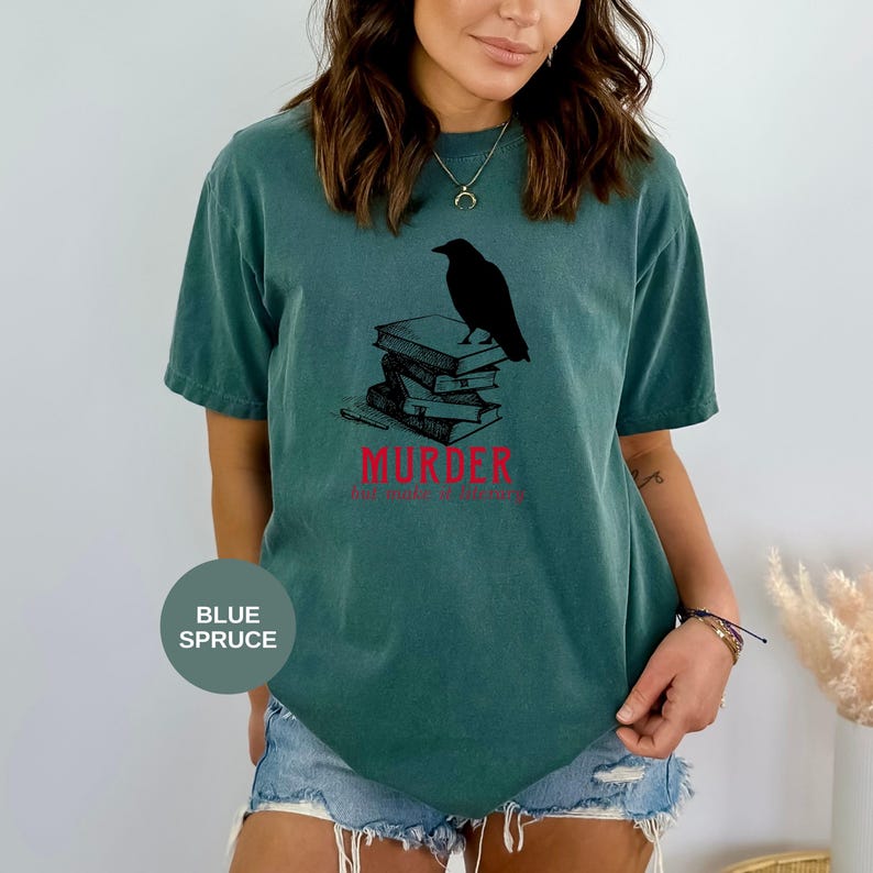 May include: Blue spruce t-shirt featuring a black raven perched atop a stack of books. The word "MURDER" is printed in red, with the phrase "her make a library" below. A casual, comfortable tee for book lovers.