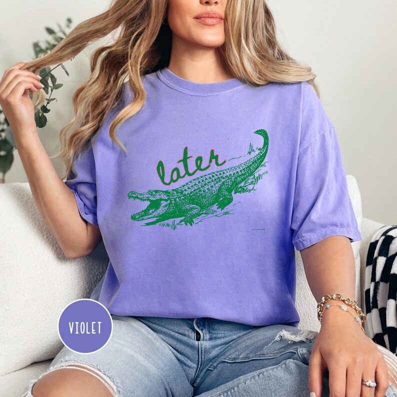 May include: Violet-colored t-shirt with a green alligator graphic and the word "later" in a script font. The shirt is a comfortable, casual style.
