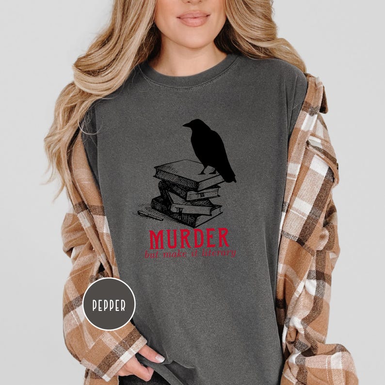 May include: Dark gray t-shirt featuring a black crow perched atop a stack of books. The text "MURDER but make it literary" is printed in red below the books. The shirt has a round neckline.