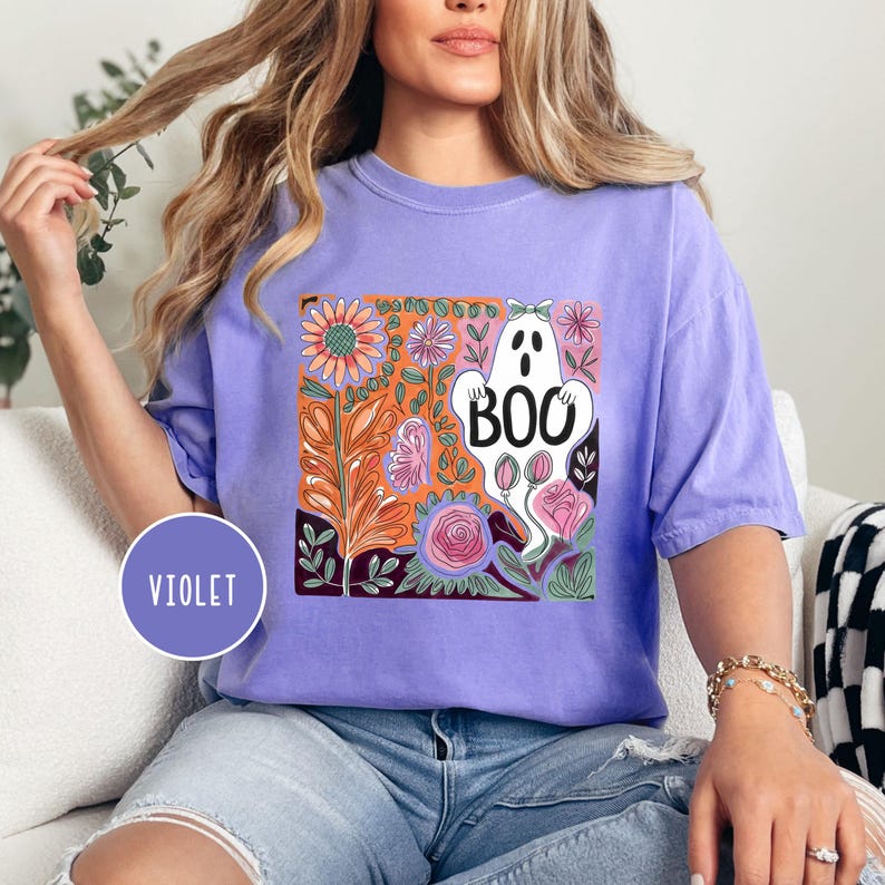 May include: A violet-colored t-shirt featuring a floral design with a white ghost that says "BOO". The design includes orange, pink, and green floral elements. The shirt has short sleeves and a relaxed fit.