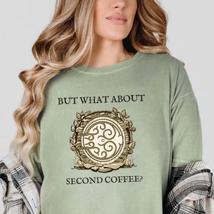 May include: Sage green t-shirt with the text "BUT WHAT ABOUT SECOND COFFEE?" above and below a circular graphic. The graphic features a wooden barrel design with an ornate, swirling center and leafy accents.