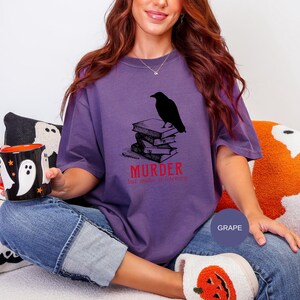 May include: A purple t-shirt with a graphic of a black raven perched on a stack of books. The word "MURDER" is printed in red below the books, with the phrase "but make it literary". The shirt is a grape color.