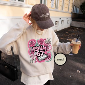May include: Sand-colored sweatshirt with a floral design featuring pink roses and the text "PSALM 67." The design includes an open book with a cross. The person is wearing a brown cap with "La Petite Paris" text.