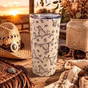 May include: A cream-colored travel tumbler with a black steer skull pattern. The tumbler has a black lid and is surrounded by a straw cowboy hat, sunglasses, and a leather bag with turquoise accents. The background features a sunset.