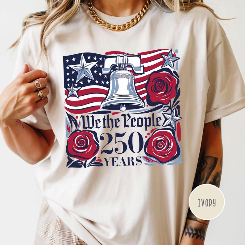 May include: Ivory-colored t-shirt featuring a patriotic design. The graphic includes the American flag, the Liberty Bell, red roses, and the text "We the People 250 Years". The design incorporates stars and stripes.