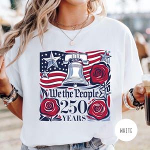 May include: White t-shirt with a patriotic design. The design includes the Liberty Bell, red roses, stars, and a stylized American flag. The text reads "We the People 250 Years."