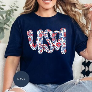 May include: A navy blue t-shirt with a white floral design that spells out "USA" in red and blue.