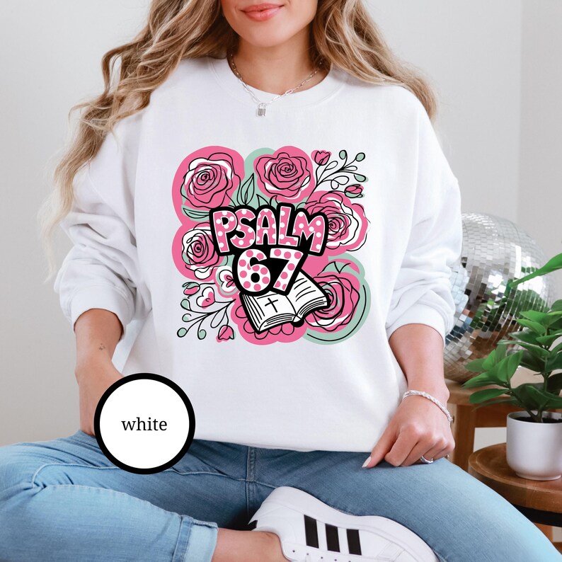 May include: White sweatshirt featuring a floral design with pink roses and the text "Psalm 67." The design includes an open book with a cross and is surrounded by green and pink accents. The word "white" is in a black circle.