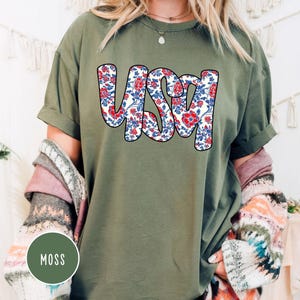 May include: A green t-shirt with a floral print of the word "USA" in red, white, and blue.