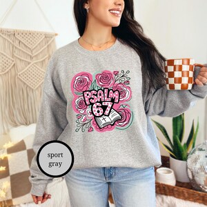 May include: Sport gray sweatshirt with a floral design and the text "PSALM 67". The design includes pink roses, a book, and the number 67. The person is holding a checkered mug.