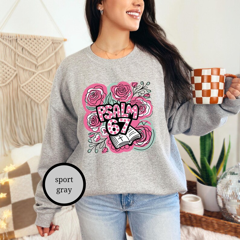 May include: Sport gray sweatshirt with a floral design and the text "PSALM 67". The design includes pink roses, a book, and the number 67. The person is holding a checkered mug.
