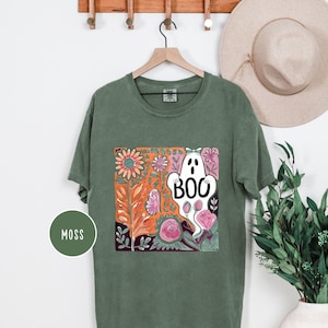 May include: Moss green t-shirt with a colorful graphic featuring a white ghost with the word "BOO" and floral elements. The design includes orange, pink, and green flowers and leaves. The shirt is hanging on a wooden hanger.