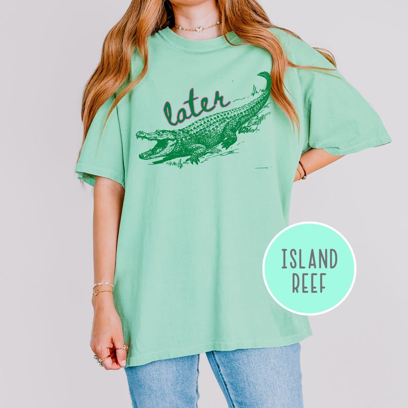 May include: A light green t-shirt with a green alligator graphic and the word "later" above it. The shirt also has a circular "Island Reef" logo on the bottom right.
