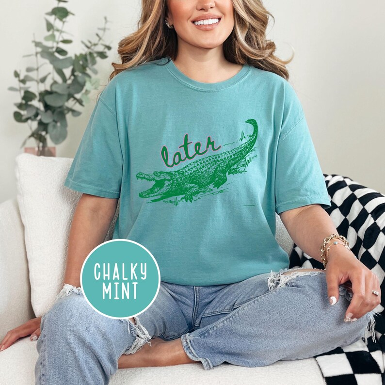 May include: Teal-colored t-shirt with a green alligator graphic and the word "later" in green script. The shirt has a relaxed fit and is made of a soft material. The shirt also has a round "Chalky Mint" logo.
