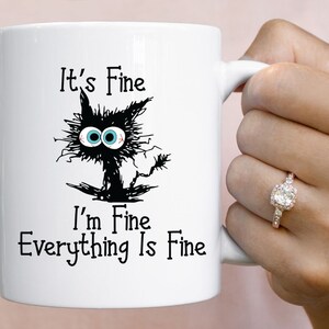 May include: White ceramic mug with a black and white illustration of a cat with the text "It's Fine I'm Fine Everything Is Fine"