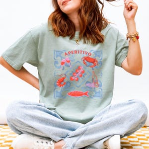 Op de afbeelding: Lichtgroen T-shirt met een blauw en roze grafisch ontwerp met het woord "APERITIVO". Het ontwerp bevat illustraties van een krab, garnalen, vissen, tomaten, wijnglazen en een fles wijn. Het shirt is gemaakt van zacht materiaal.