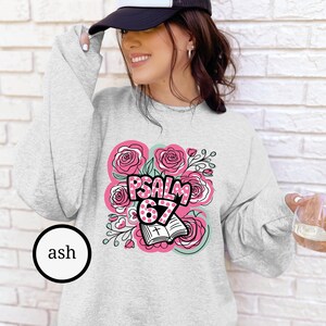 May include: Ash-colored sweatshirt featuring a floral design with pink roses and the text "PSALM 67". The design includes an open book with a cross. The sweatshirt has long sleeves and a crew neck.