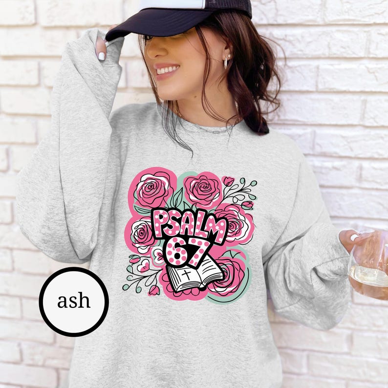 May include: Ash-colored sweatshirt featuring a floral design with pink roses and the text "PSALM 67". The design includes an open book with a cross. The sweatshirt has long sleeves and a crew neck.