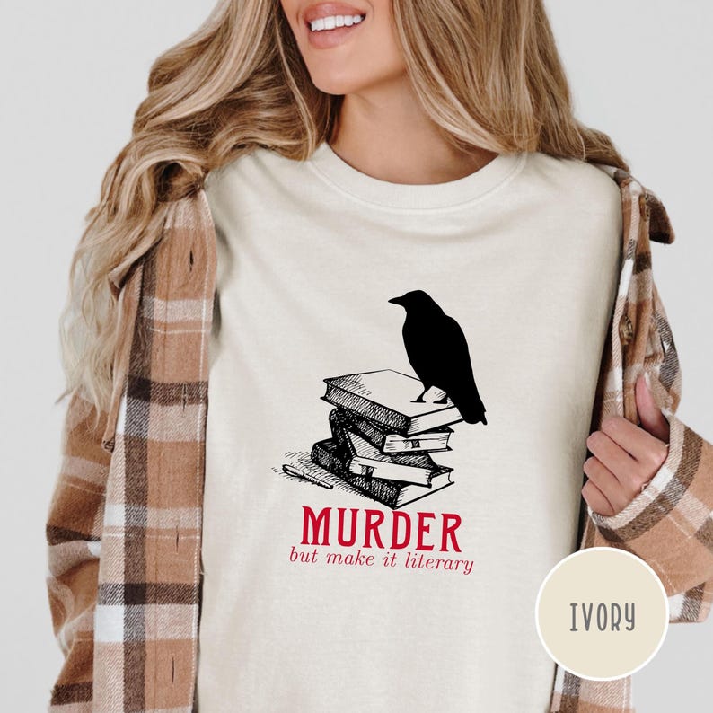 May include: Ivory-colored t-shirt featuring a black crow perched atop a stack of books. The text "MURDER but make it literary" is printed in red. The shirt has a crew neck and short sleeves.