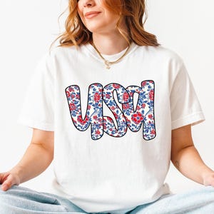 May include: White t-shirt with a red, white, and blue floral pattern spelling out "USA".