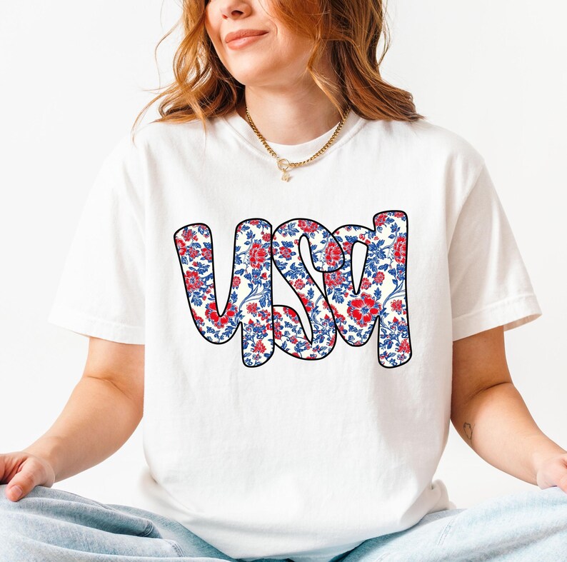 May include: White t-shirt with a red, white, and blue floral pattern spelling out "USA".